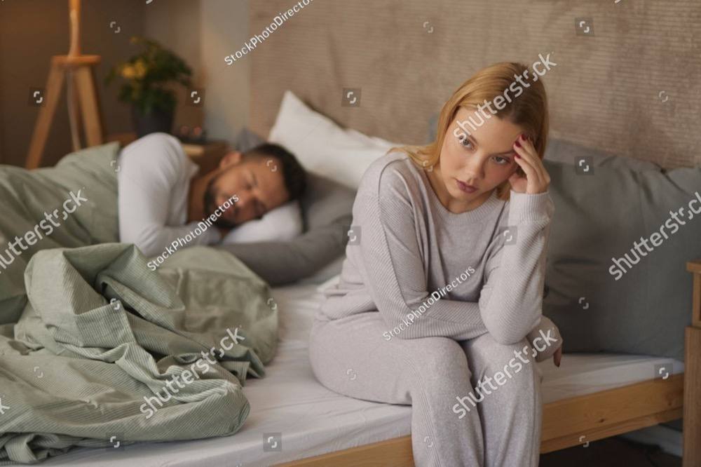 A woman sits on the edge of the bed at dawn, her partner asleep and turned away behind her