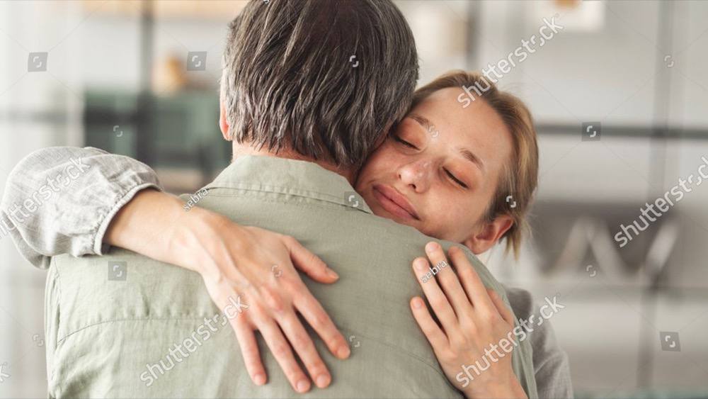 An adult daughter holds her older father, eyes closed